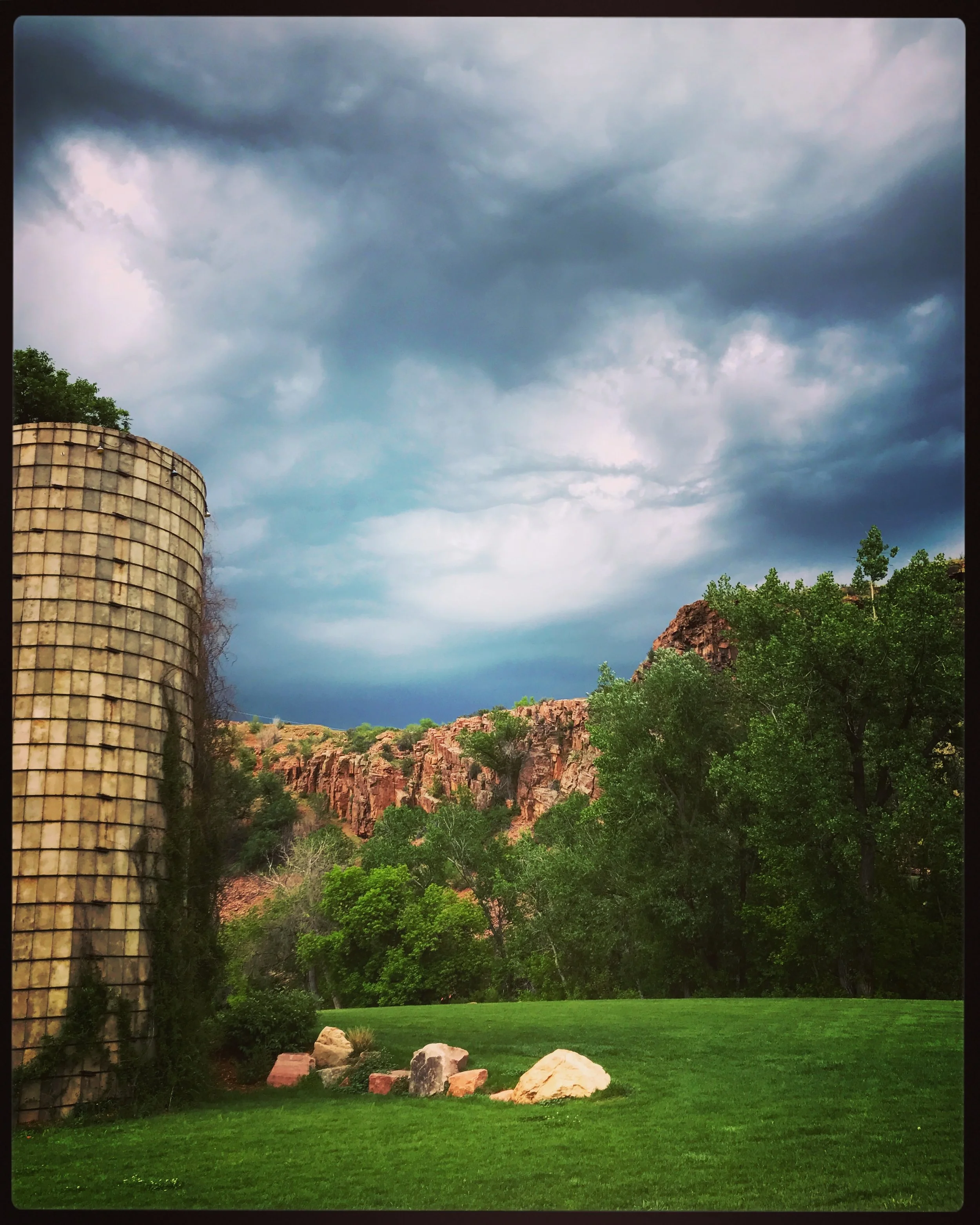 A landscape scene featuring a dramatic, cloudy sky over red rocks and green trees, with a grassy area and rocks in the foreground, and part of an old farm silo on the left side at Planet Bluegrass Ranch in Lyons, Colorado.