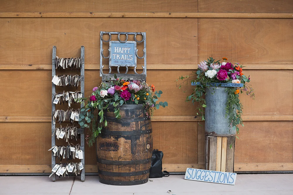 Decorative display with flower arrangements, a barrel, a rustic ladder with tags, and a sign that says 'Happy Trails' against a wooden wall at a wedding at Planet Bluegrass.