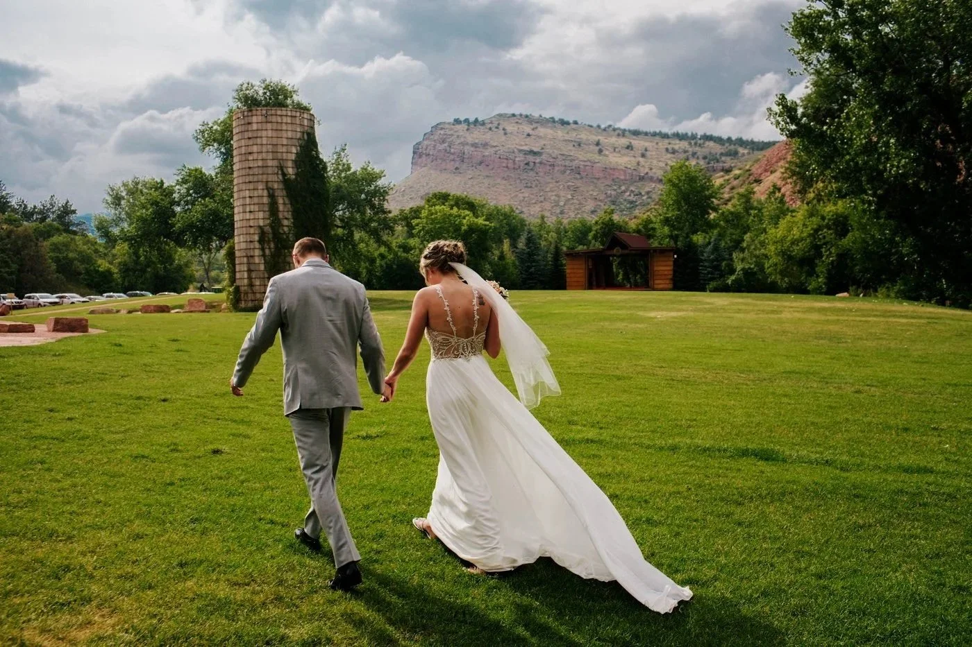 A bride and groom walk hand in hand across a grassy field on their wedding day, with a scenic mountain and cloudy sky in the background.