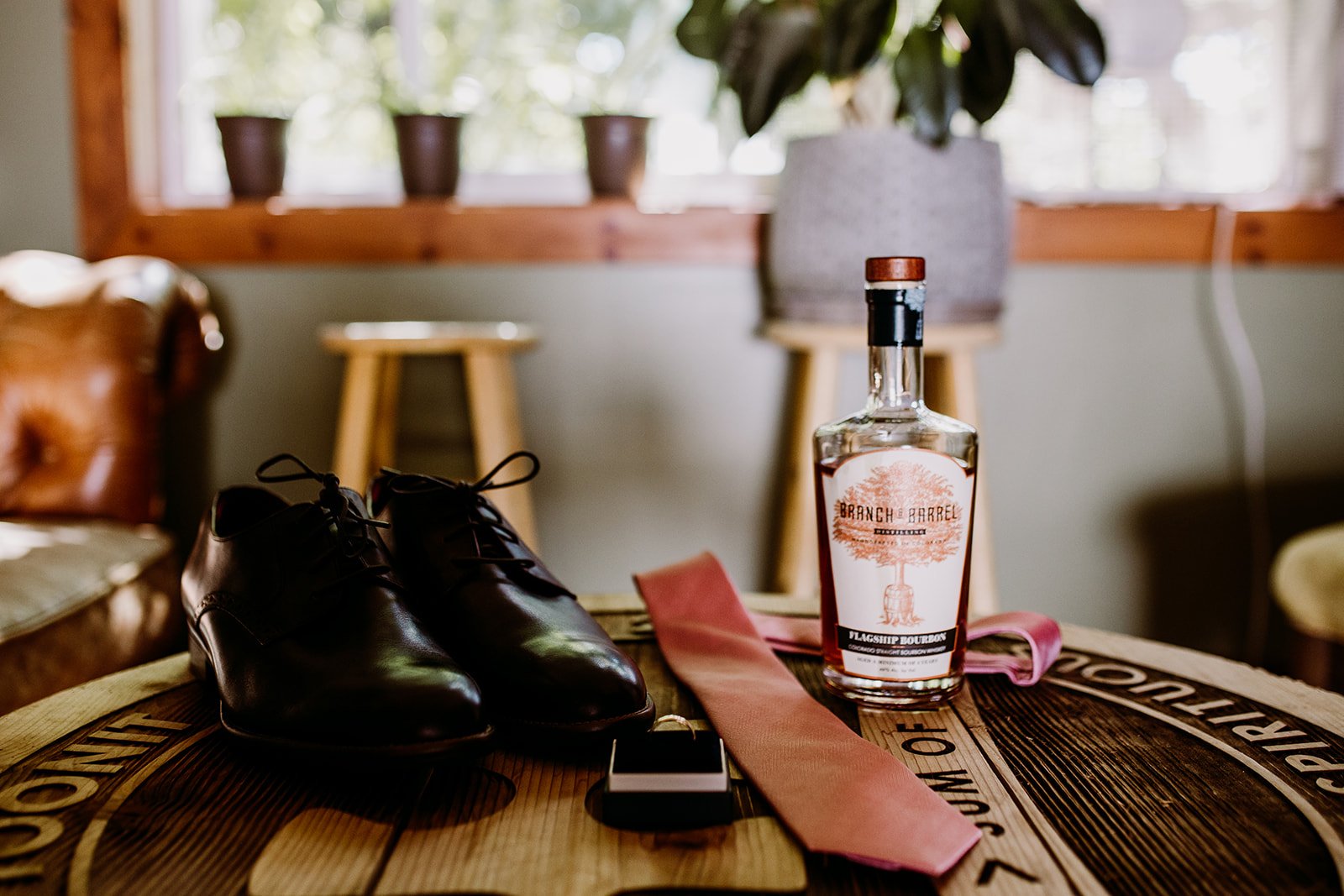 Pair of black leather dress shoes, a bottle of bourbon labeled 'Barn & Barrel Flagship Bourbon,' a pink cloth, and a smartphone on a wooden barrel table inside a cozy room with a window and potted plants in the background.