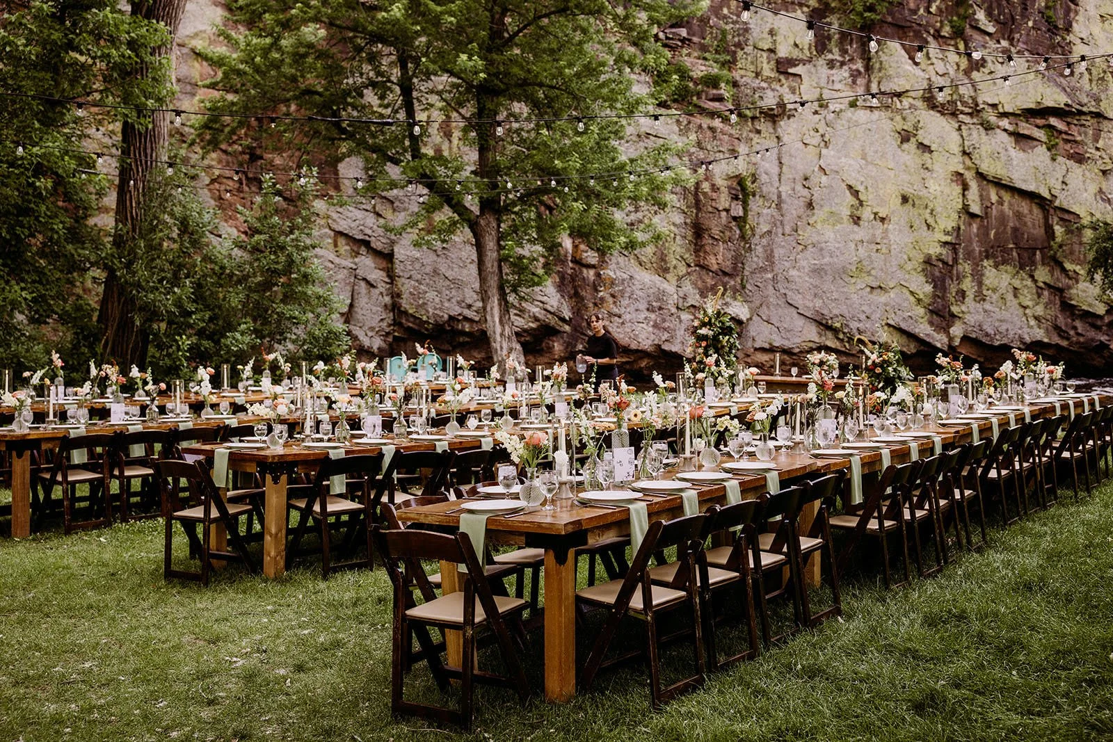 Outdoor wedding setup at Planet Bluegrass with long wooden tables and chairs, decorated with flowers, glassware, and candles, on a grassy area with a rocky cliff and trees in the background, and string lights overhead.
