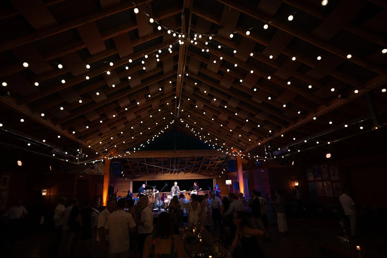 An indoor event with a live band performing on stage, surrounded by people dancing and socializing. The ceiling is decorated with string lights.