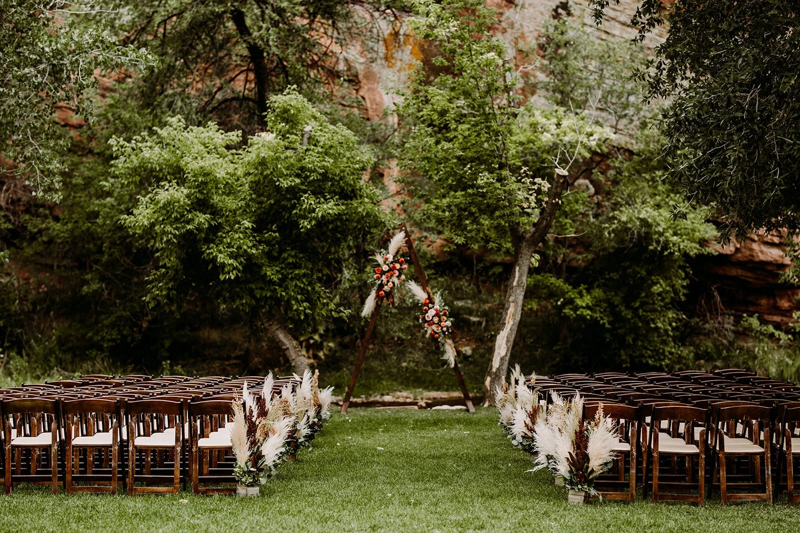 Outdoor wedding ceremony setup in a grassy area with rows of wooden chairs on either side of a green aisle, leading to a triangular floral arch decorated with pink flowers and white pampas grass, surrounded by trees and natural rock formations.