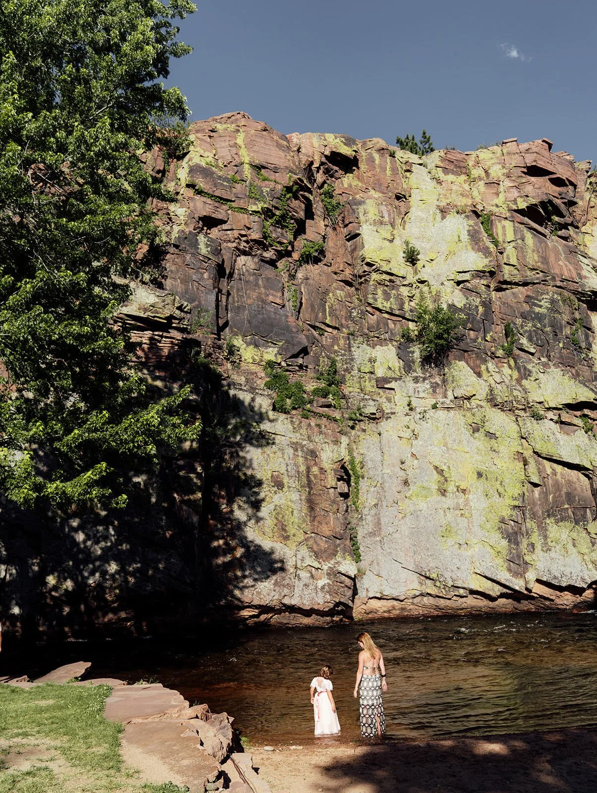 Wedding guests, a woman and a young girl, standing in a river near a large rocky cliff at Planet Bluegrass with trees and clear blue sky.