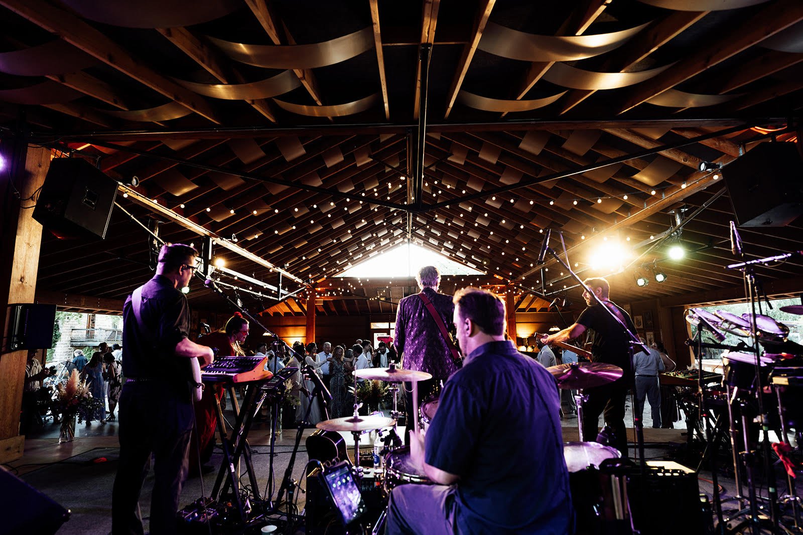 A live band performing on stage inside a venue with an audience attending, under a wooden ceiling with string lights and bright stage lighting.