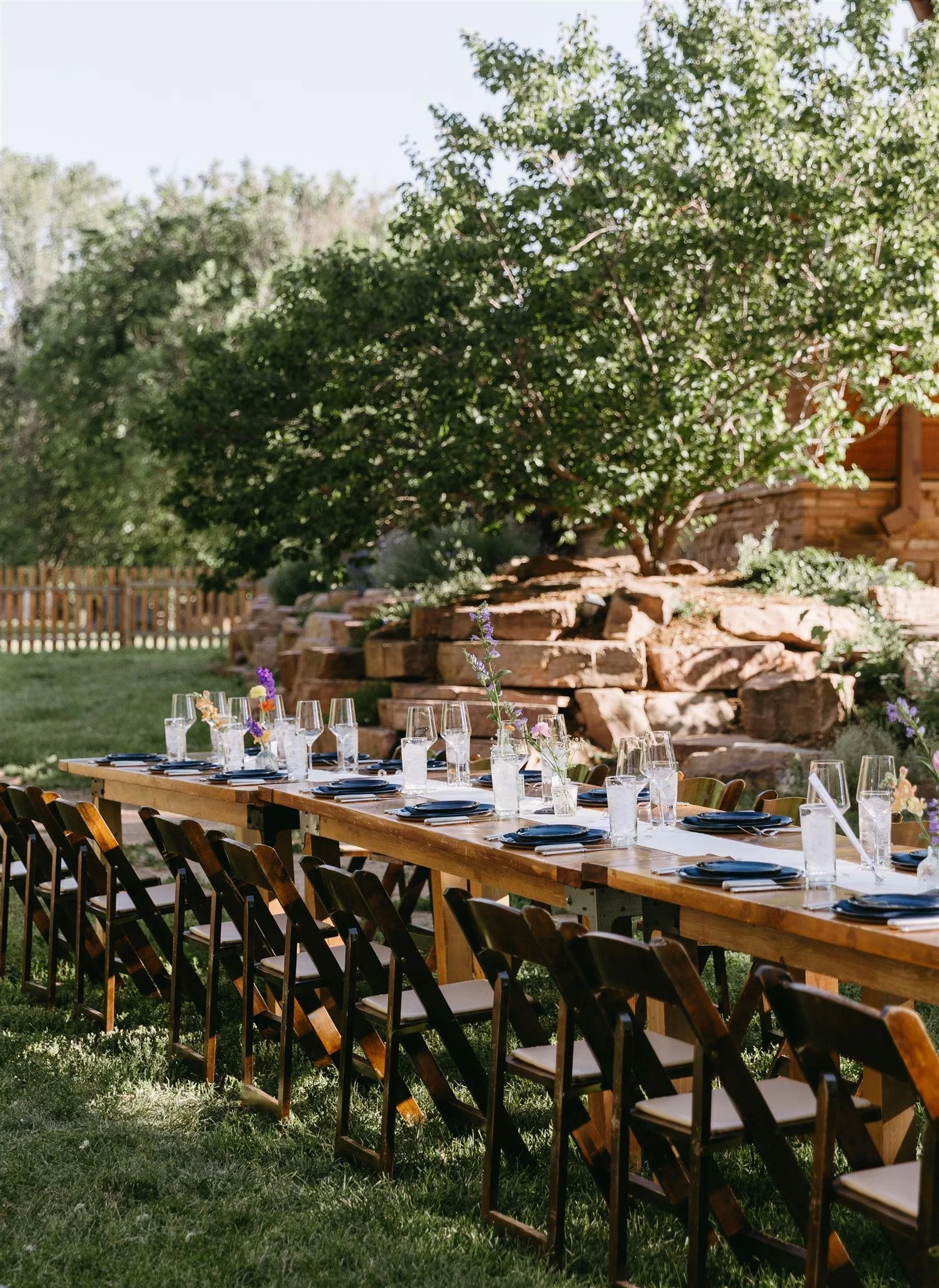 Long wooden dining table set for a wedding with blue plates, wine glasses, and small flower arrangements, outdoors in a grassy field at Planet Bluegrass Ranch with a large tree and stone steps in the background.