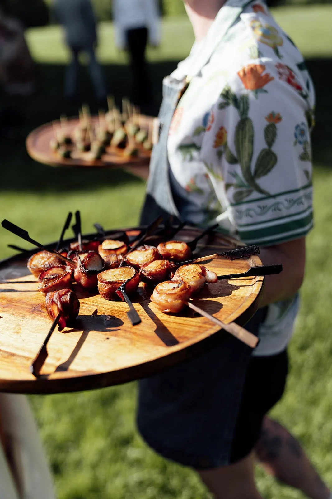 Person holding a wooden tray with grilled meat skewers outdoors during the daytime, blurred green background, wearing a floral shirt and dark shorts.