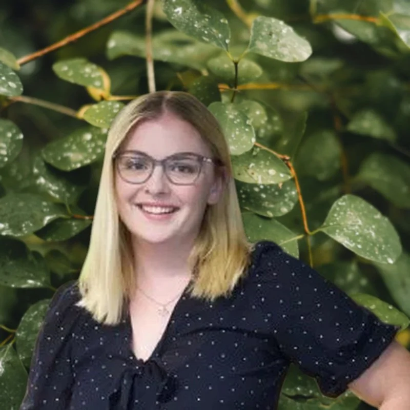 A smiling woman with blonde hair and glasses standing in front of a large green leafy plant background.