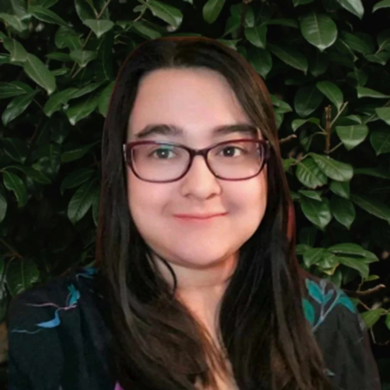 A woman with glasses and long dark hair standing in front of leafy green plants.