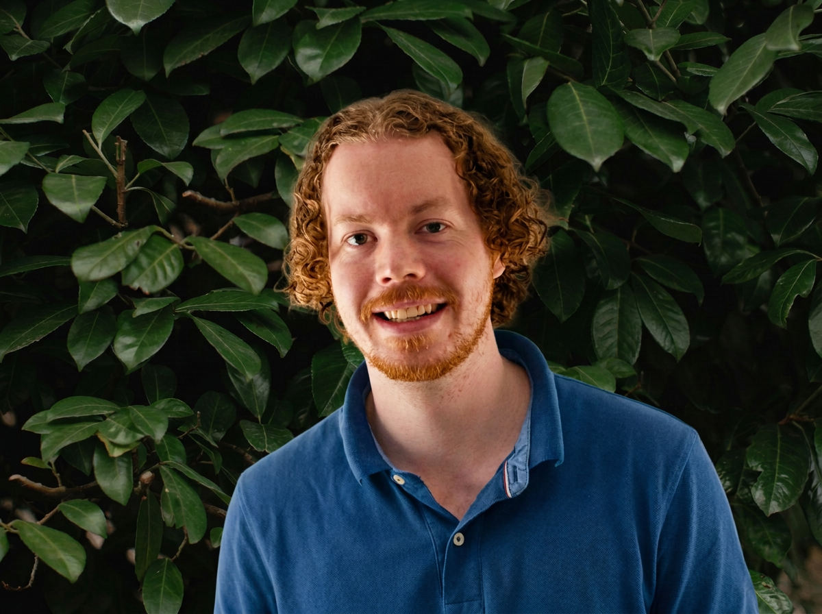 A young man with curly red hair and a beard, wearing a blue polo shirt, standing in front of lush green foliage and smiling.