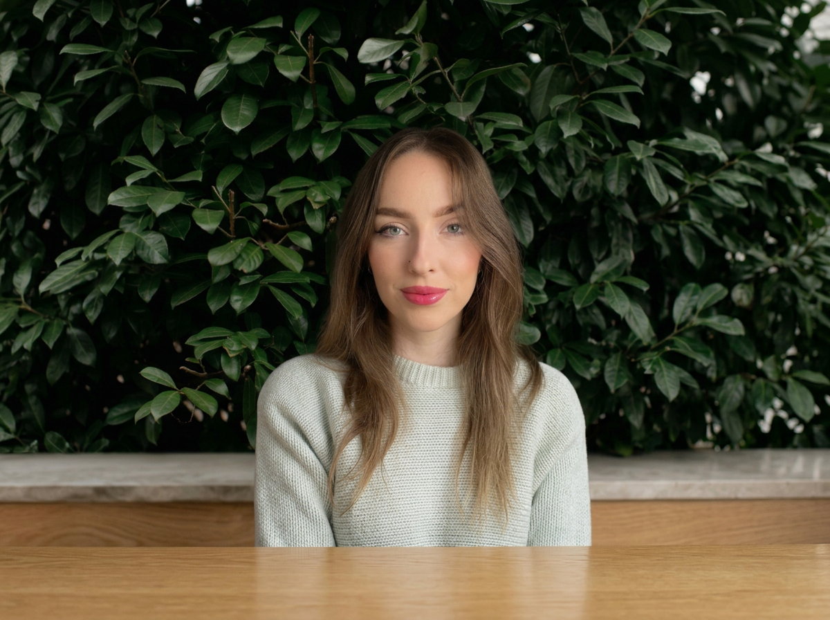 A young woman with long, wavy brown hair and blue eyes, wearing a light gray sweater, sitting in front of a lush green leafy background.