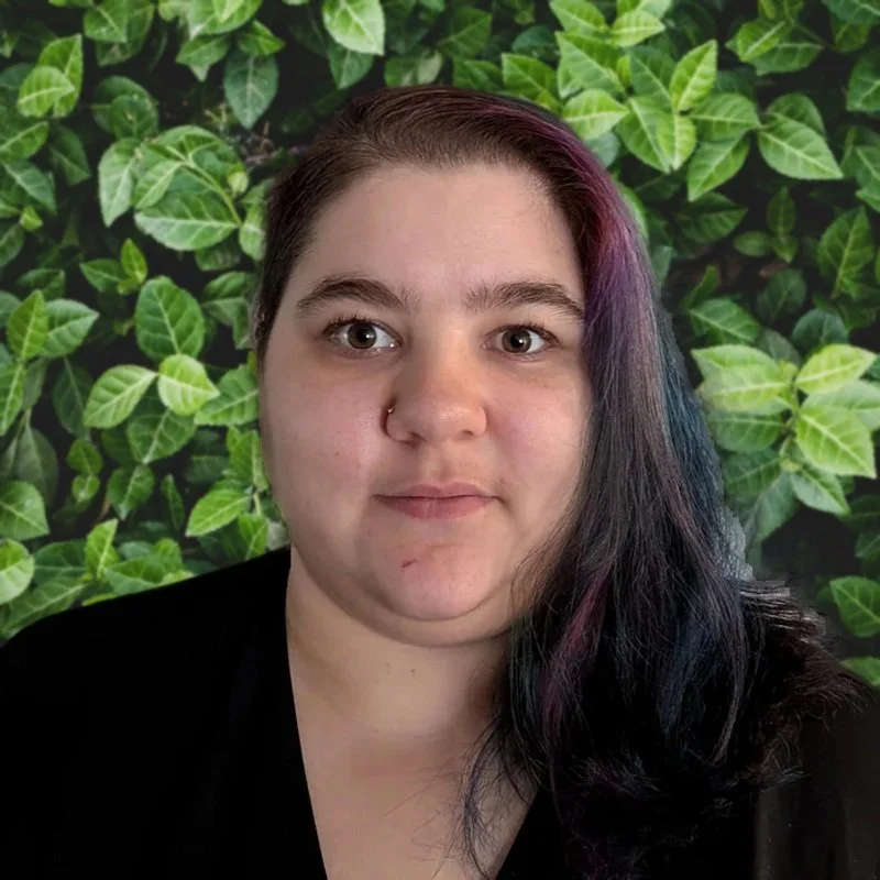 A woman with dark hair that has streaks of purple and blue, wearing a black top, in front of a background filled with green leafy plants.