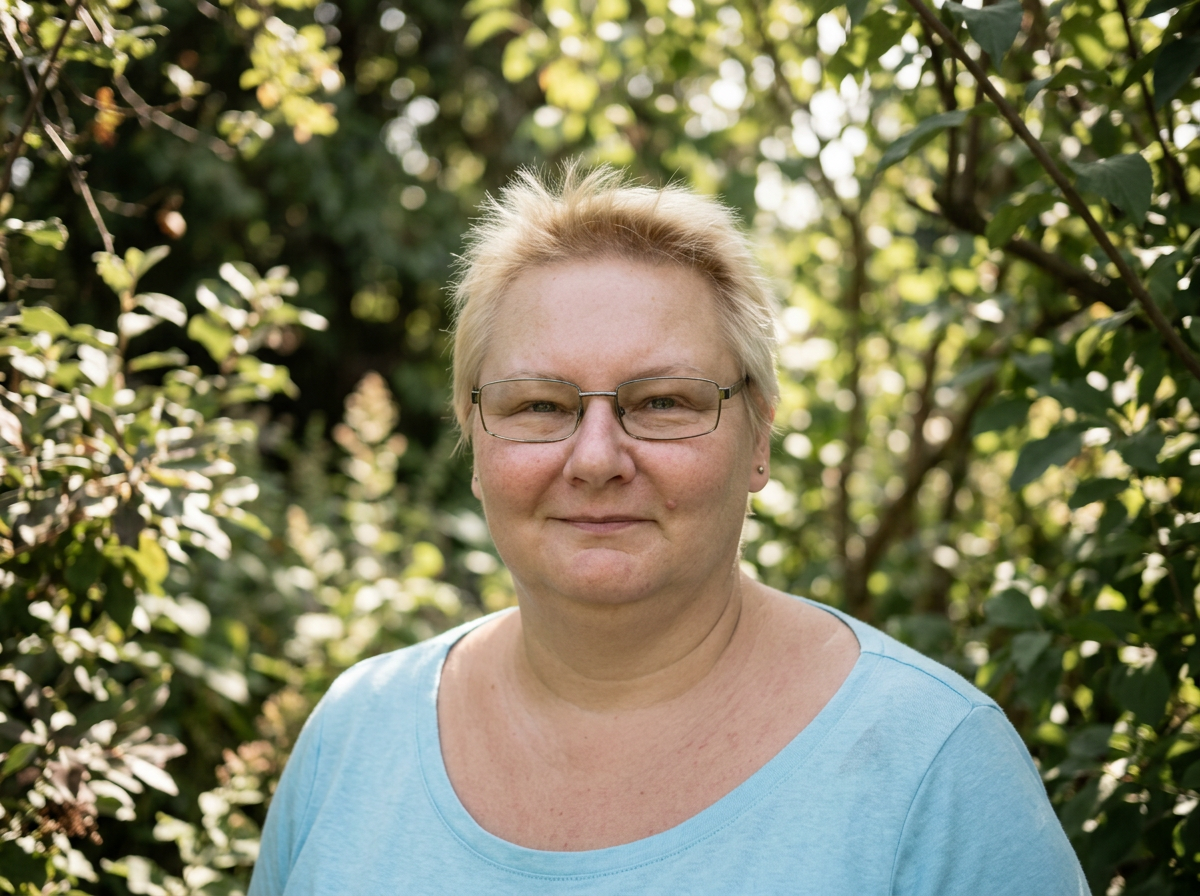 A woman with short blonde hair and glasses standing outdoors in front of lush green foliage, wearing a light blue shirt.