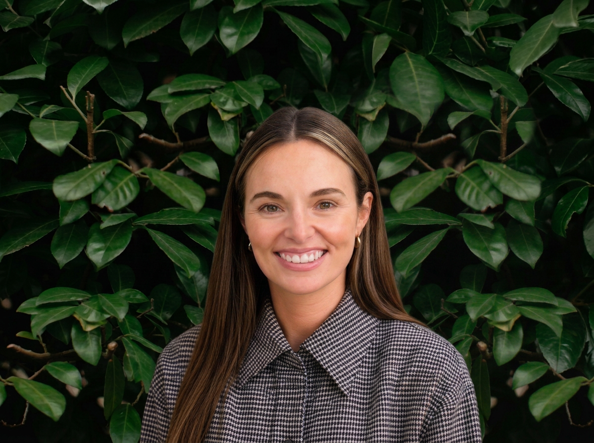 A woman with long brown hair smiling, wearing a checkered shirt, standing in front of lush green leafy bushes.