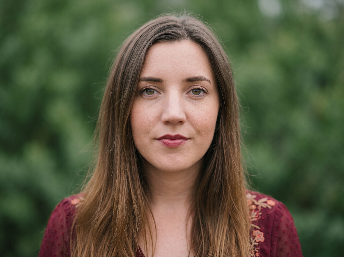 A woman with long brown hair and green eyes standing outdoors with a blurred green background.