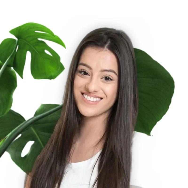 A young woman smiling with long dark hair, standing in front of a background of large green monstera leaves.