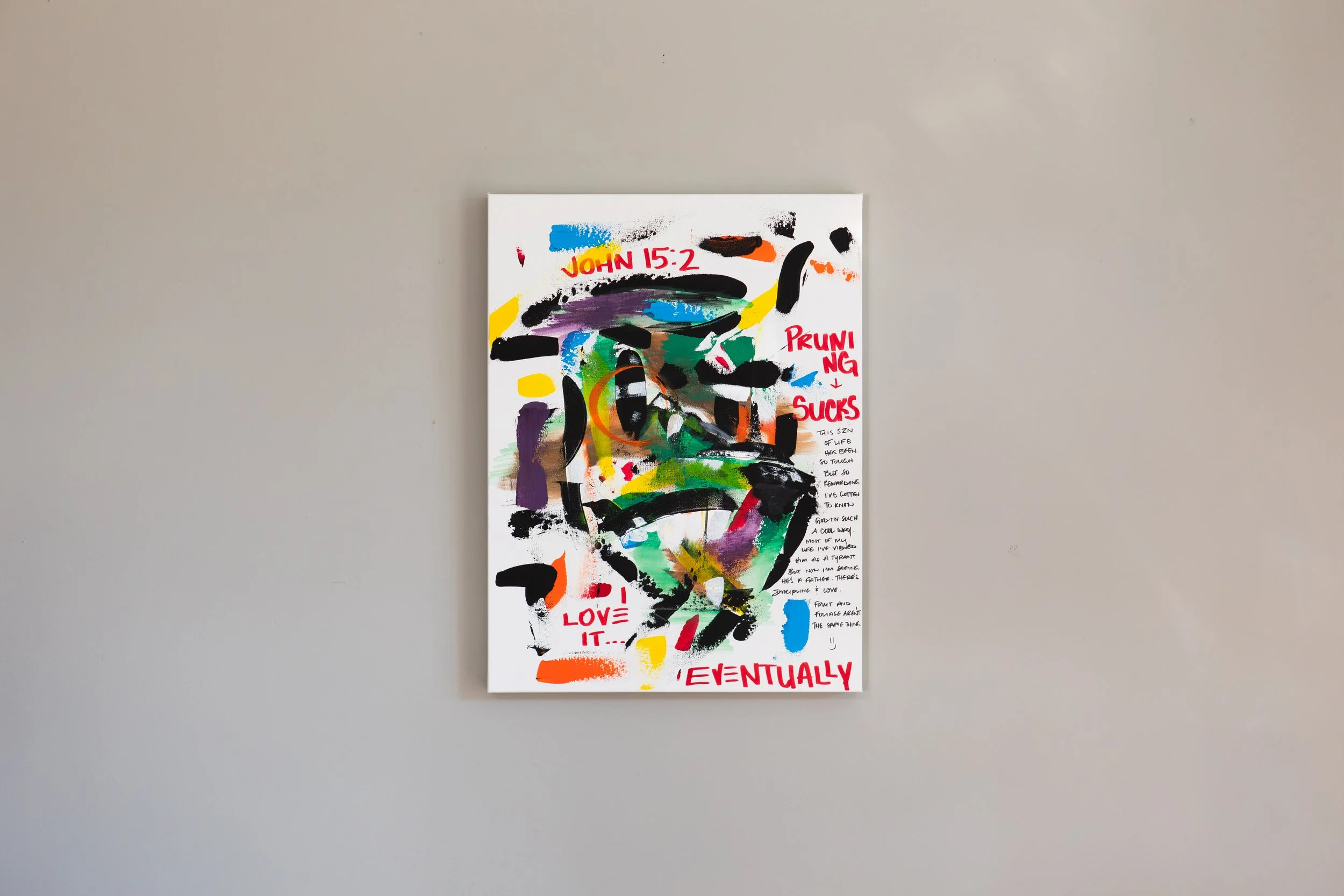 Colorful abstract painting with splashes of black, blue, yellow, green, purple, and orange on a white canvas, with handwritten text in red and black, including phrases like 'JOHN 15:2', 'PRUNING SUCKS', 'LOVE IT', and 'EVENTUALLY' on a plain beige wall.