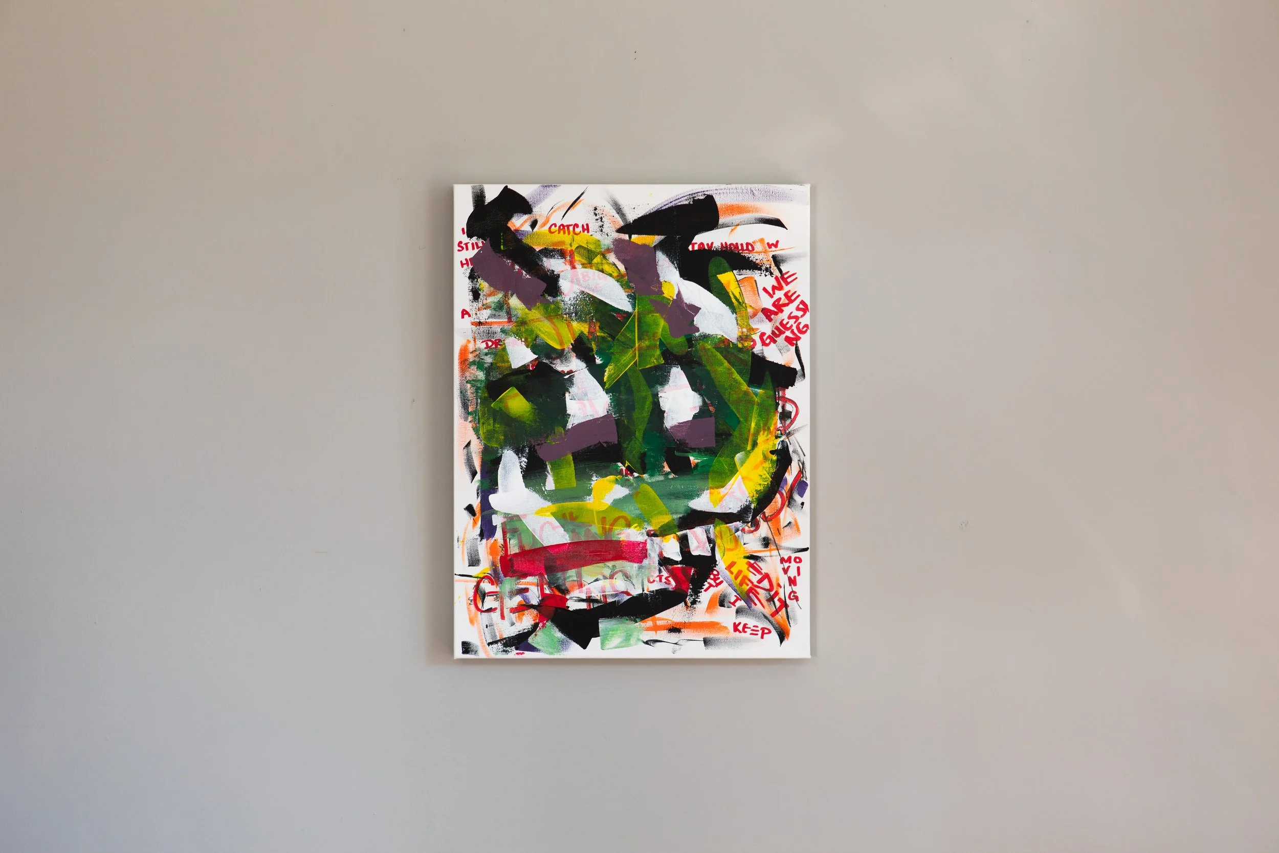 Abstract colorful painting with black, green, purple, red, and orange brushstrokes and red text on a white background, hung on a plain light gray wall.