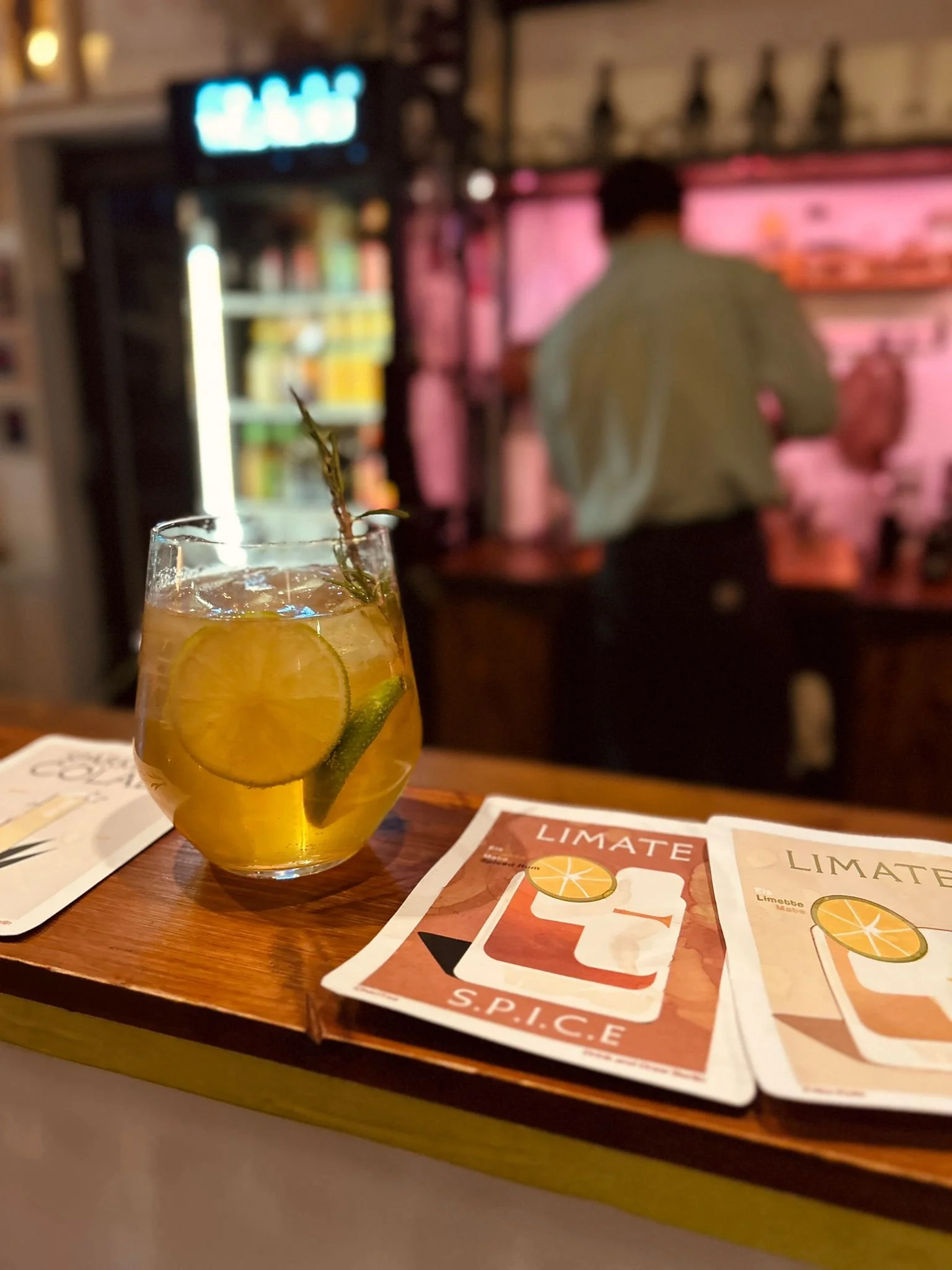 Tasty looking cocktail long drink on a wooden bar with a cozy lit bar space and bartender at drink and draw berlin atelier studio space