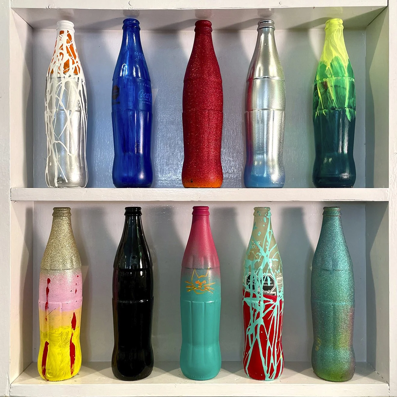  Media Litre 10 #2   10 painted 17 oz Mexican Coke bottles in a custom-built , gloss-white painted wooden wall display. Dimensions: 24” x 24” x 3.5”  