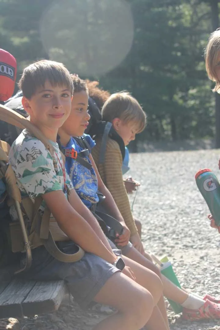 A camper with an overnight camping backpack smiles while looking at the camera at Camp Arrowhead for boys. There are two other camper friends on the bench looking forward.