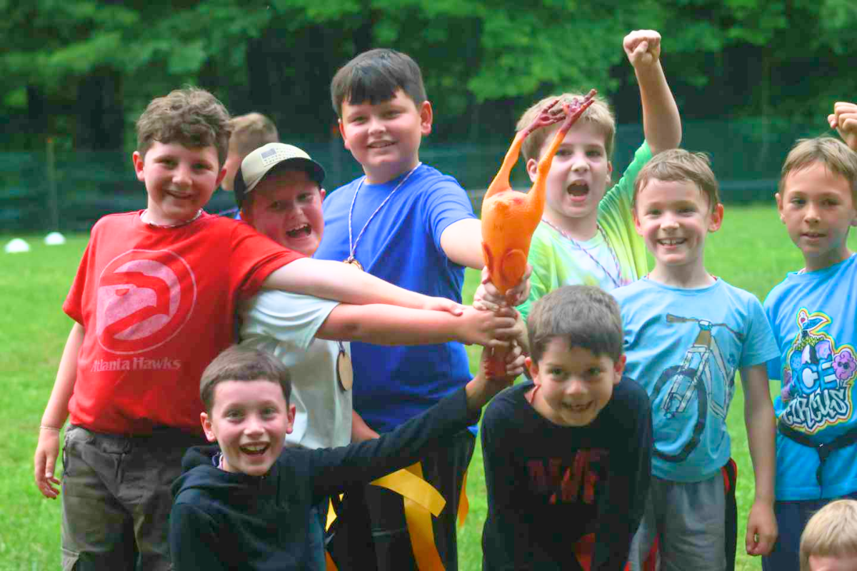 A group of younger overnight campers play a camp favorite game on the field at Camp Arrowhead for boys. They're smiling while holding a rubber chicken