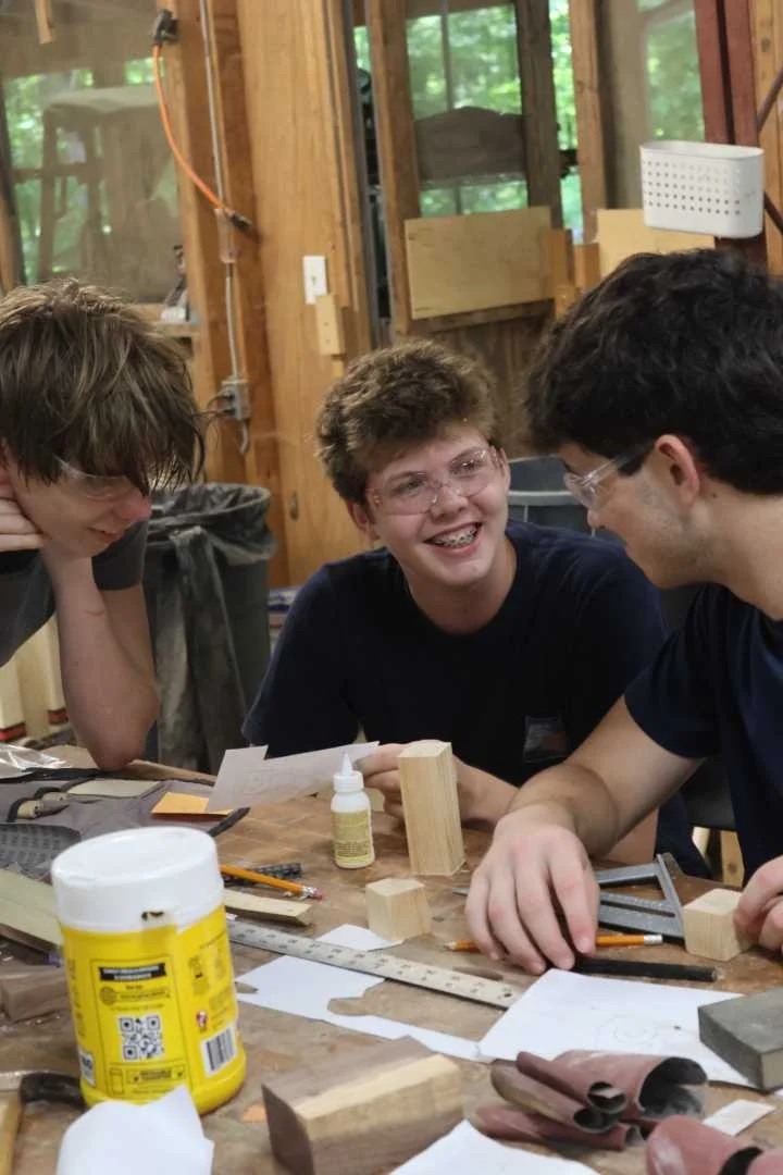 A happy teen camper is smiling with friends while in the woodworking shop at Camp Arrowhead for boys.