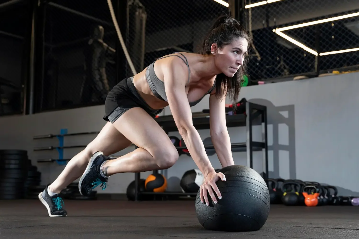 A woman working out in a gym: on her hands and feet, pushing a black medicine ball while wearing a gray sports bra and black shorts.