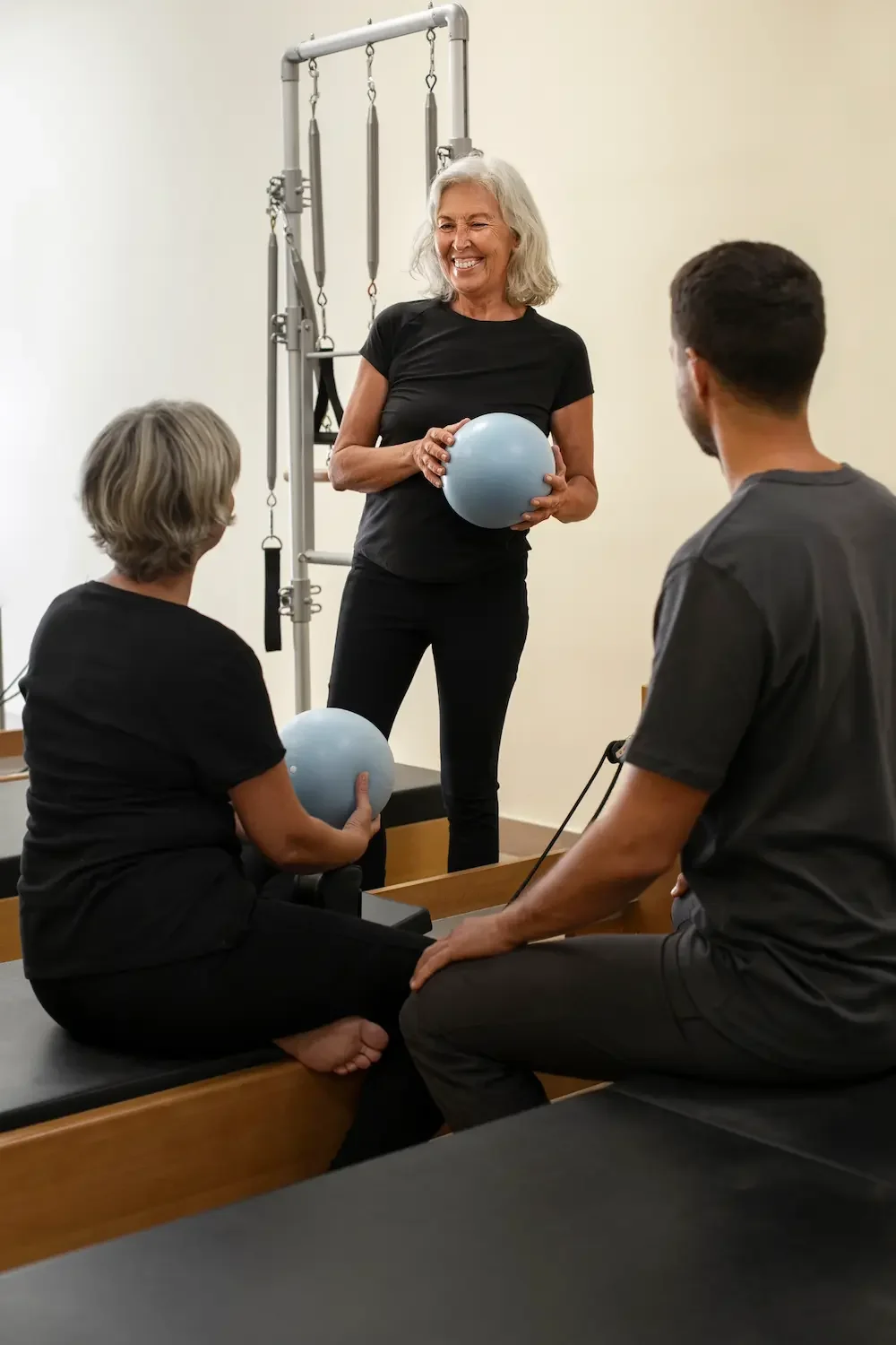 An elderly woman holding a light blue exercise ball, standing in a physical therapy or rehabilitation setting. She is smiling and appears to be instructing two younger adults, one holding a similar exercise ball, while they sit on black therapy tables.