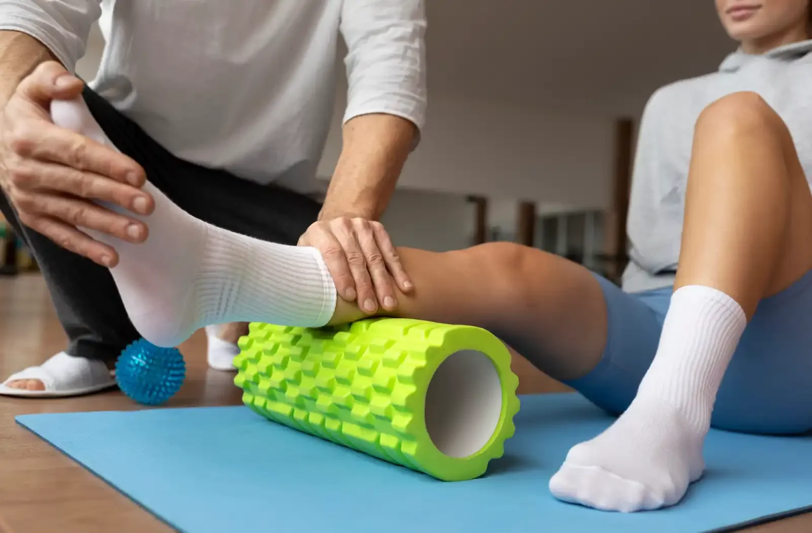 Physical therapy session with a person using a foam roller on the leg, assisted by a therapist, on a blue yoga mat.