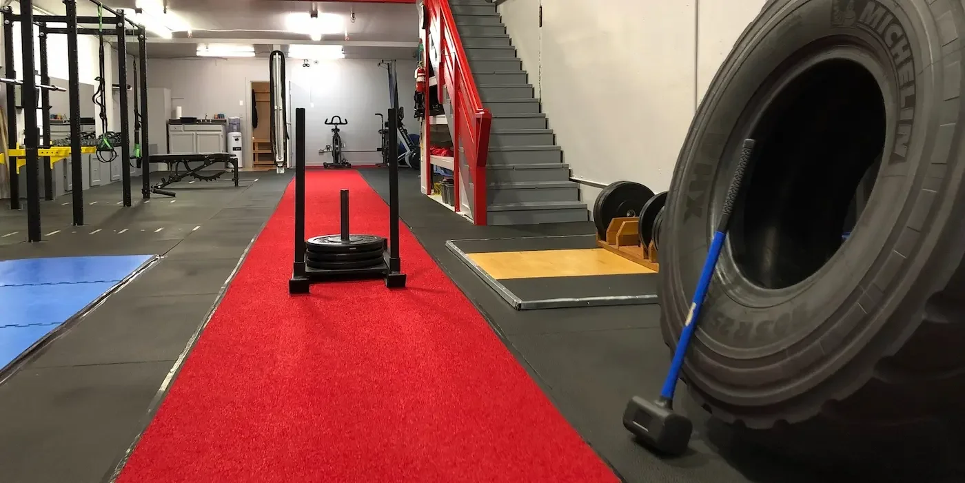 Inside a gym with various fitness equipment including weight plates, a tire, a sled, and a red carpeted area for exercises.