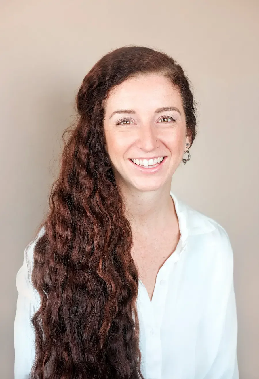 A young woman with long, wavy red hair smiling, wearing a white blouse, against a plain beige background.
