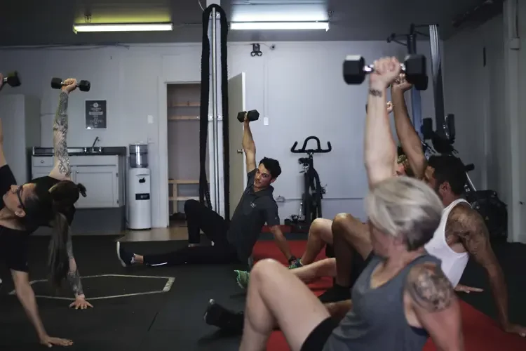 People exercise with dumbbells in a gym, seated and lying on mats, in a group workout session.