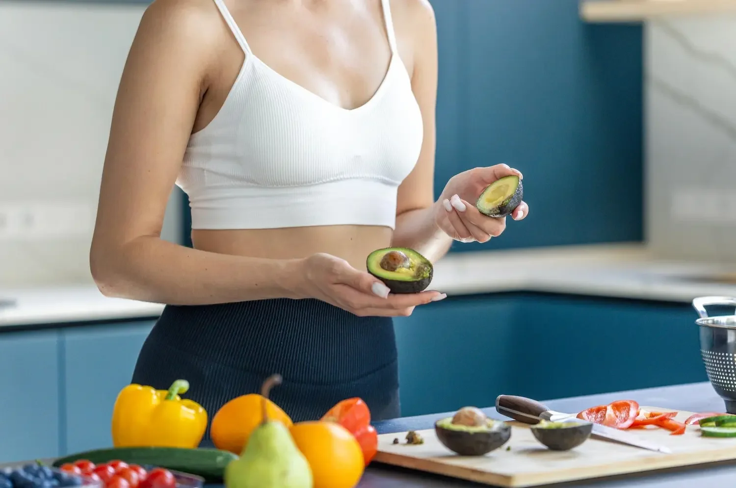 A person in a white crop top and dark pants holding an avocado cut in half in a modern kitchen with colorful vegetables on the counter.