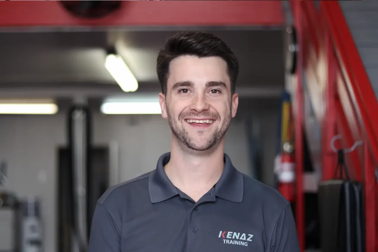 A smiling young man with dark hair and a beard, wearing a dark gray polo shirt with 'Kenaz Training' logo, standing inside a gym or training center with exercise equipment visible in the background.