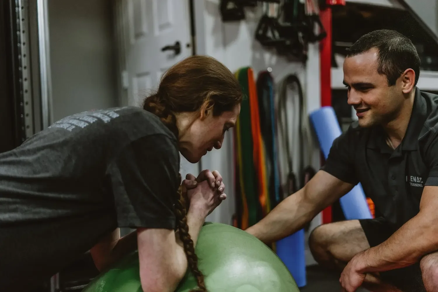 A woman doing a plank exercise with her elbows resting on a green exercise ball, being supported and encouraged by a trainer in a gym.