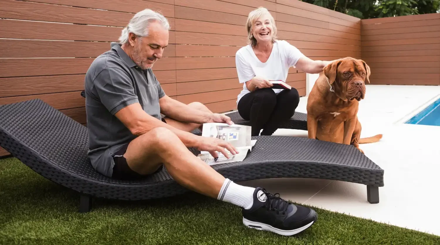 An elderly man with gray hair and a beard relaxing on a black outdoor lounge chair, reading a magazine, next to an elderly woman with gray hair, smiling, sitting on the ground with a notebook on her lap, petting a large brown dog sitting by her side near a pool, wooden fence in the background.