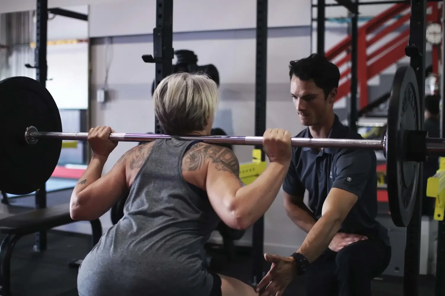 A woman with short gray hair and tattoos on her arm is doing a squat with a barbell, while a fitness trainer is providing support and guidance at a gym.
