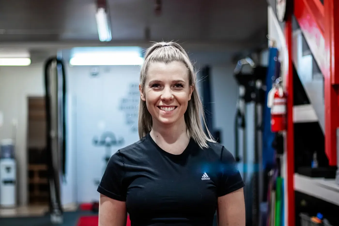 A smiling woman with blonde hair tied in a high ponytail standing in a gym surrounded by workout equipment.