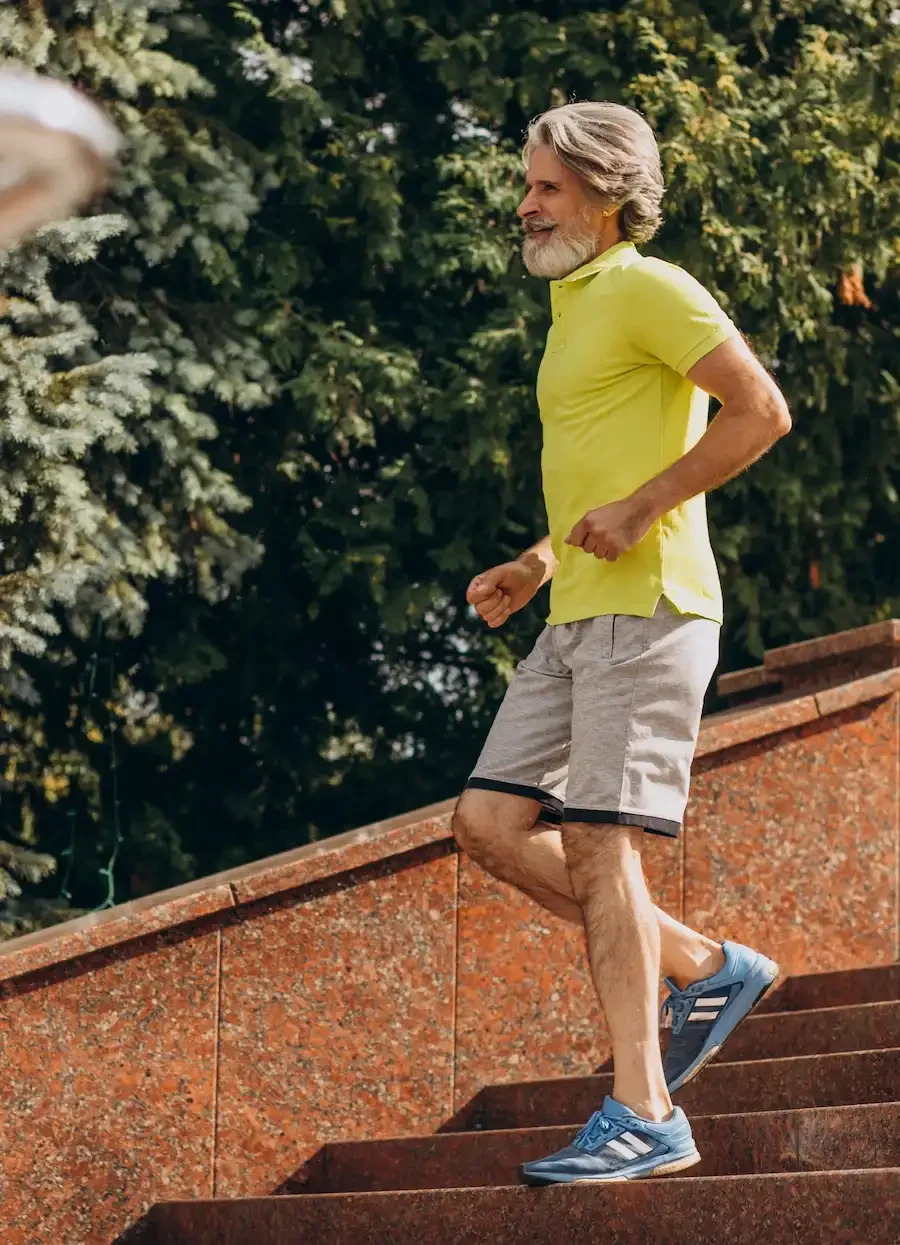 An older man with gray hair and a beard, wearing a yellow polo shirt, light gray shorts, and blue running shoes, jogging up outdoor stairs. Green trees are in the background.
