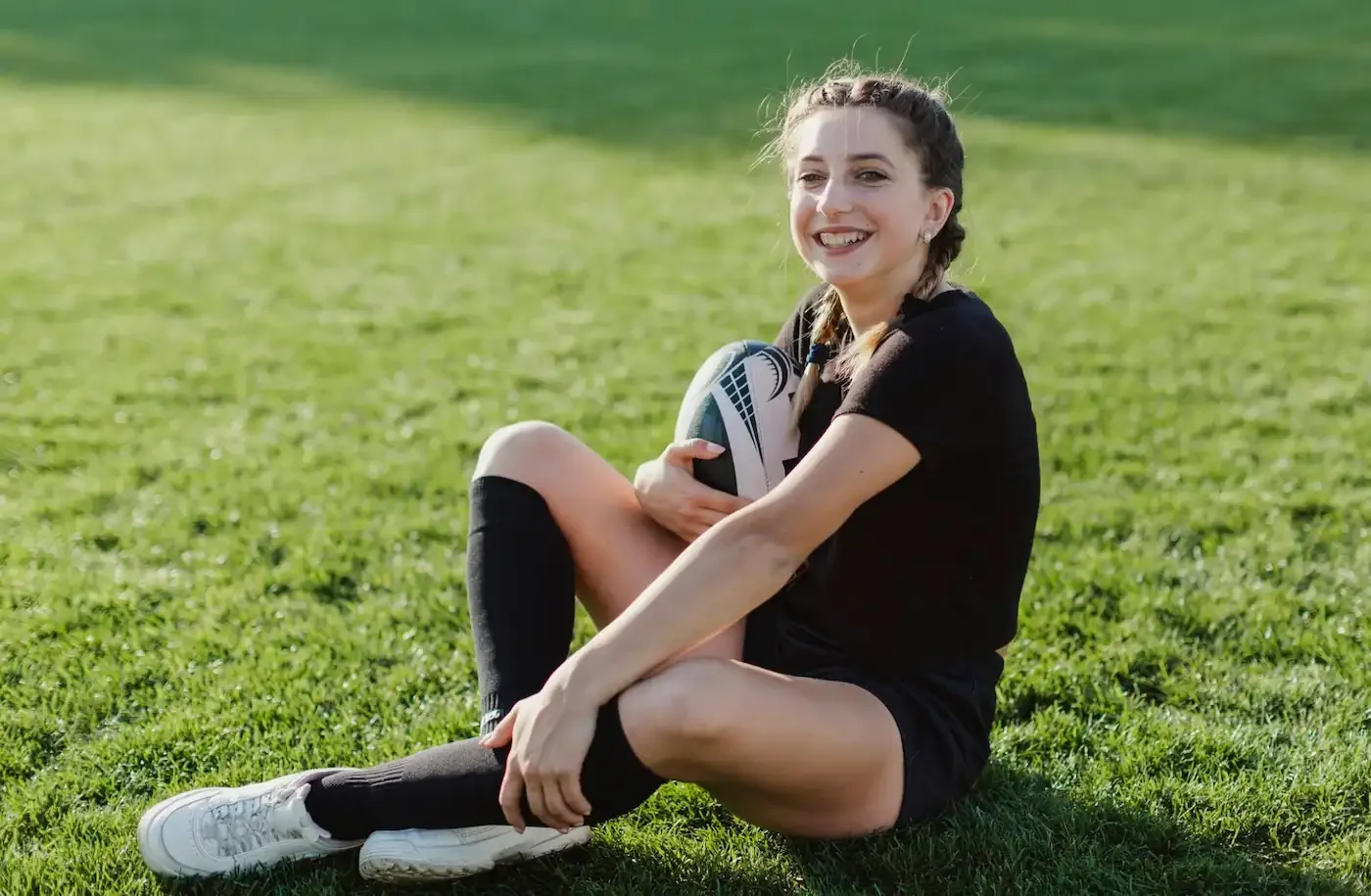 Young woman with a rugby ball sitting on grass field, smiling at camera, wearing black sports outfit and knee-high socks.