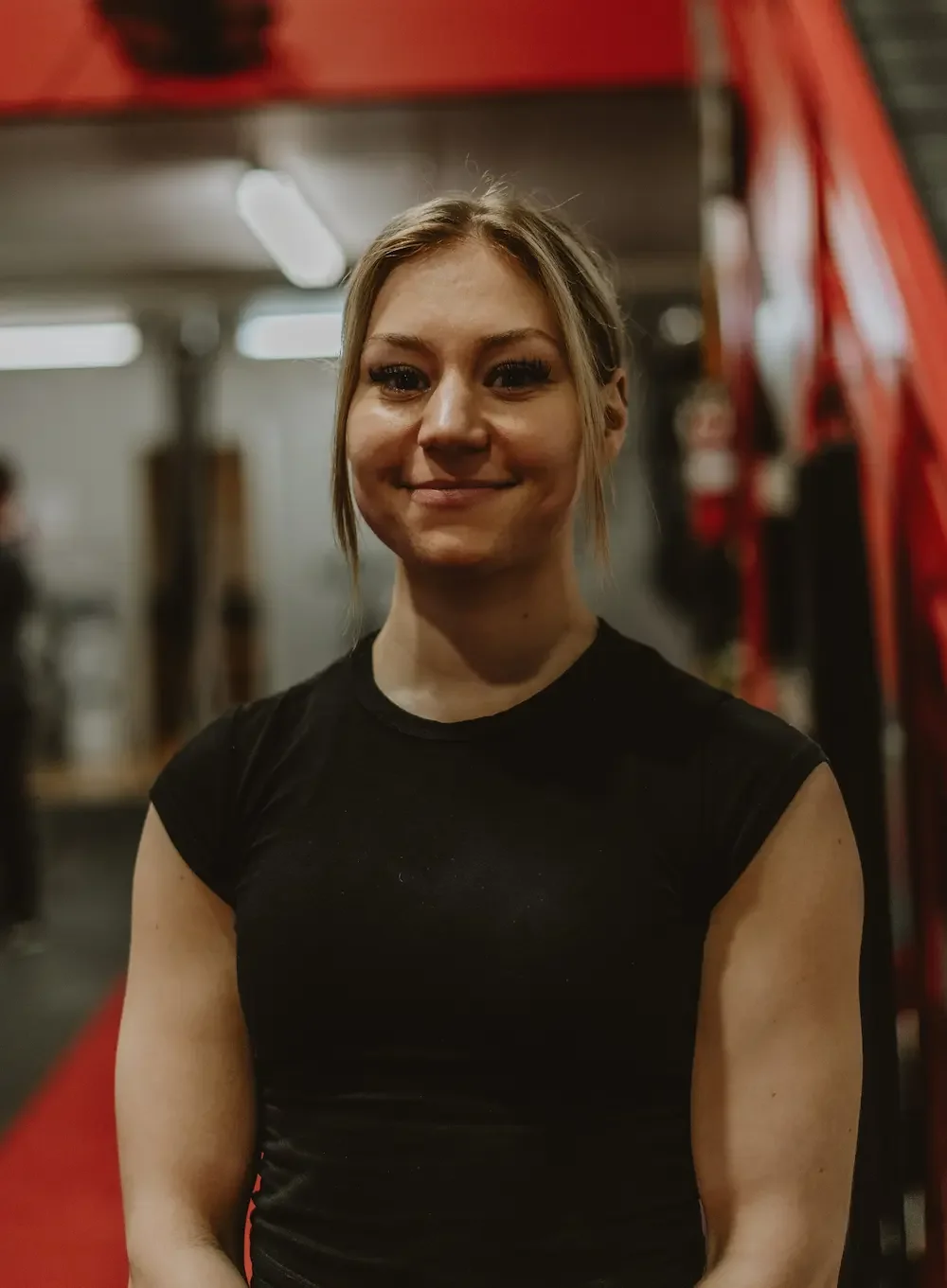 A young woman with blonde hair and a black sleeveless shirt smiling indoors, against a background of gym equipment and red structural elements.