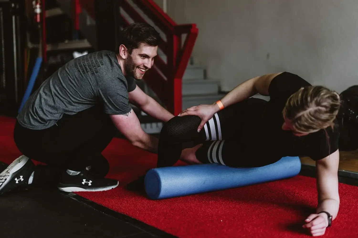 A trainer assisting a woman in doing a workout on a foam roller at the gym, with both smiling.