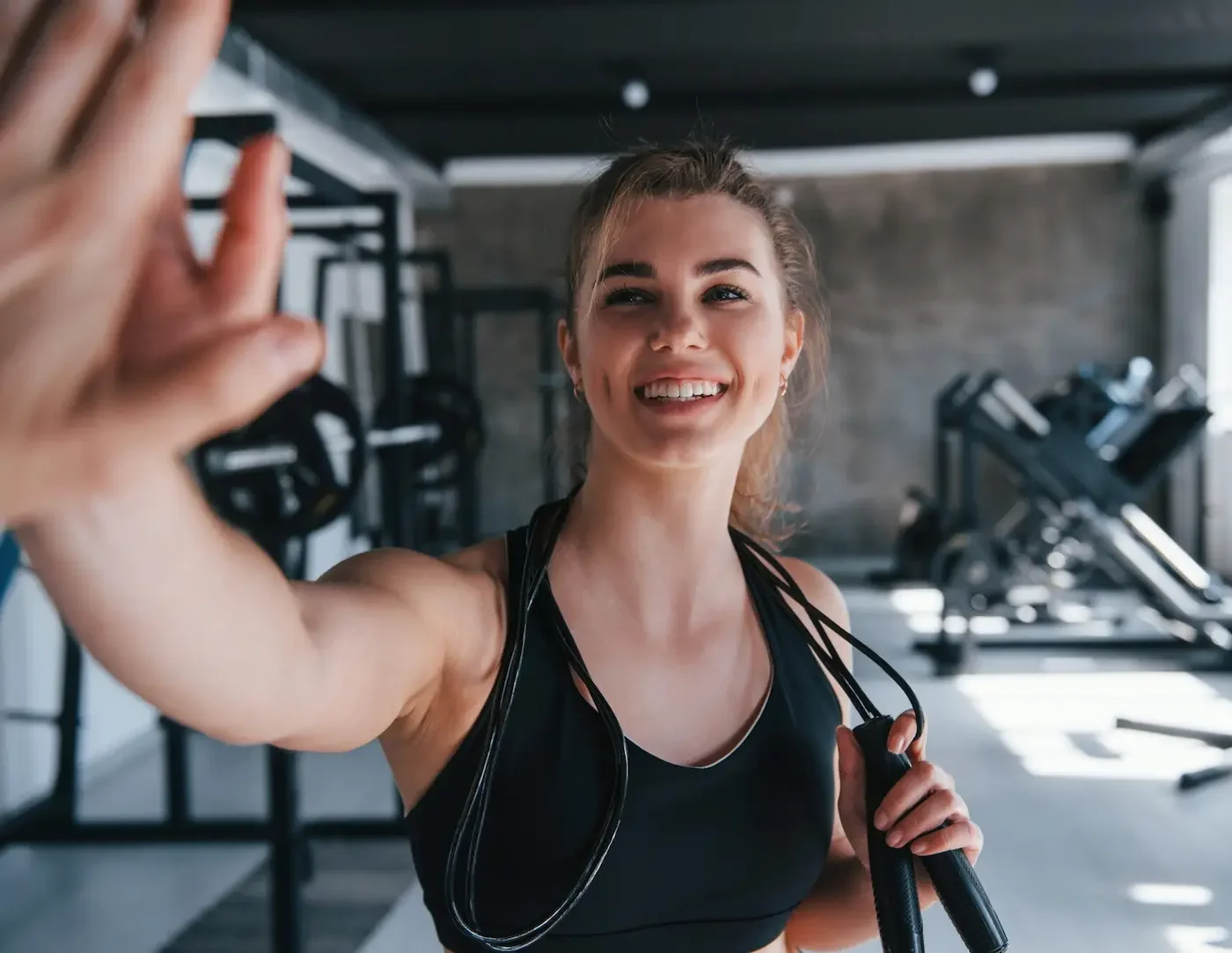 A young woman smiling and taking a selfie at the gym, holding a jump rope, with workout equipment in the background.