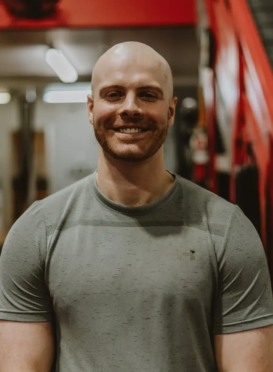 A smiling man with a shaved head and beard, wearing a gray athletic shirt, standing in a gym.