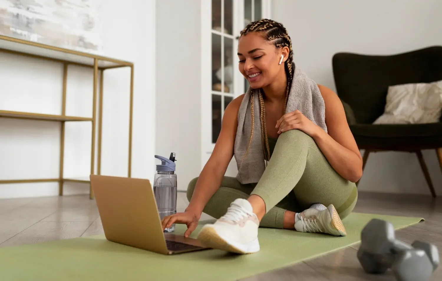 A woman in athletic wear sitting on a yoga mat, smiling, with a laptop, water bottle, and dumbbells in a home gym or living space.
