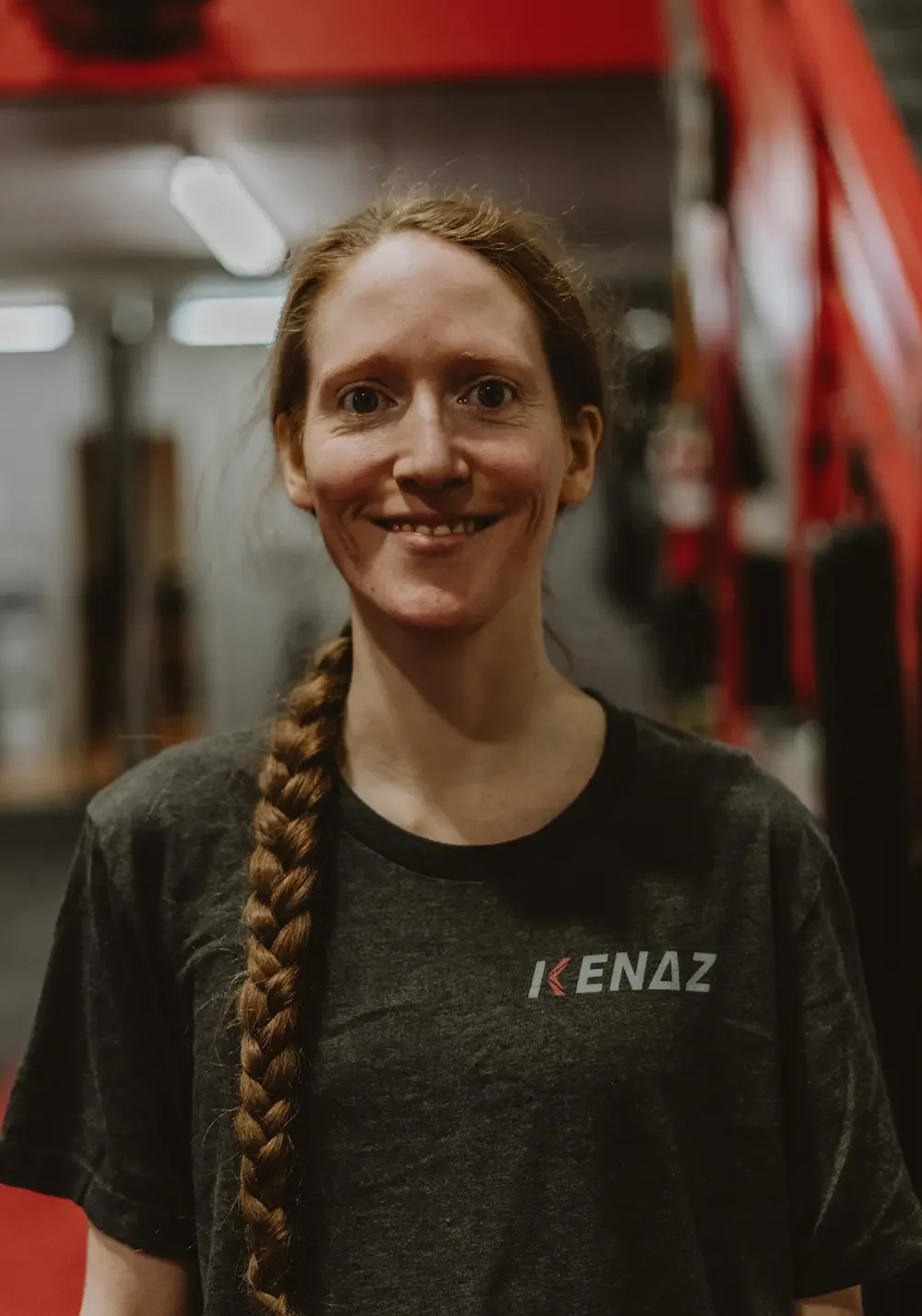 A woman with long braided hair smiling in a gym, wearing a black T-shirt with the 'KENAZ' logo.