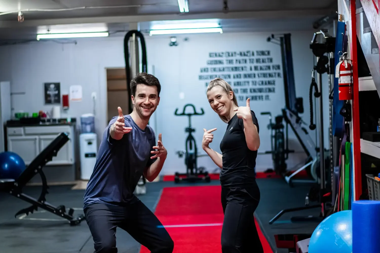 A man and woman smiling and posing in a gym, pointing at each other, surrounded by exercise equipment like kettlebells, stability balls, and resistance bands.