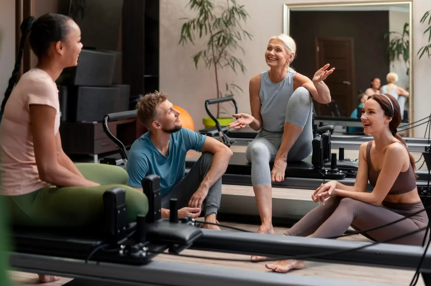 Group of women and a man in a fitness studio, engaged in conversation during a workout session. One woman is standing, and the others are sitting or kneeling on the floor or on exercise equipment, smiling and listening.