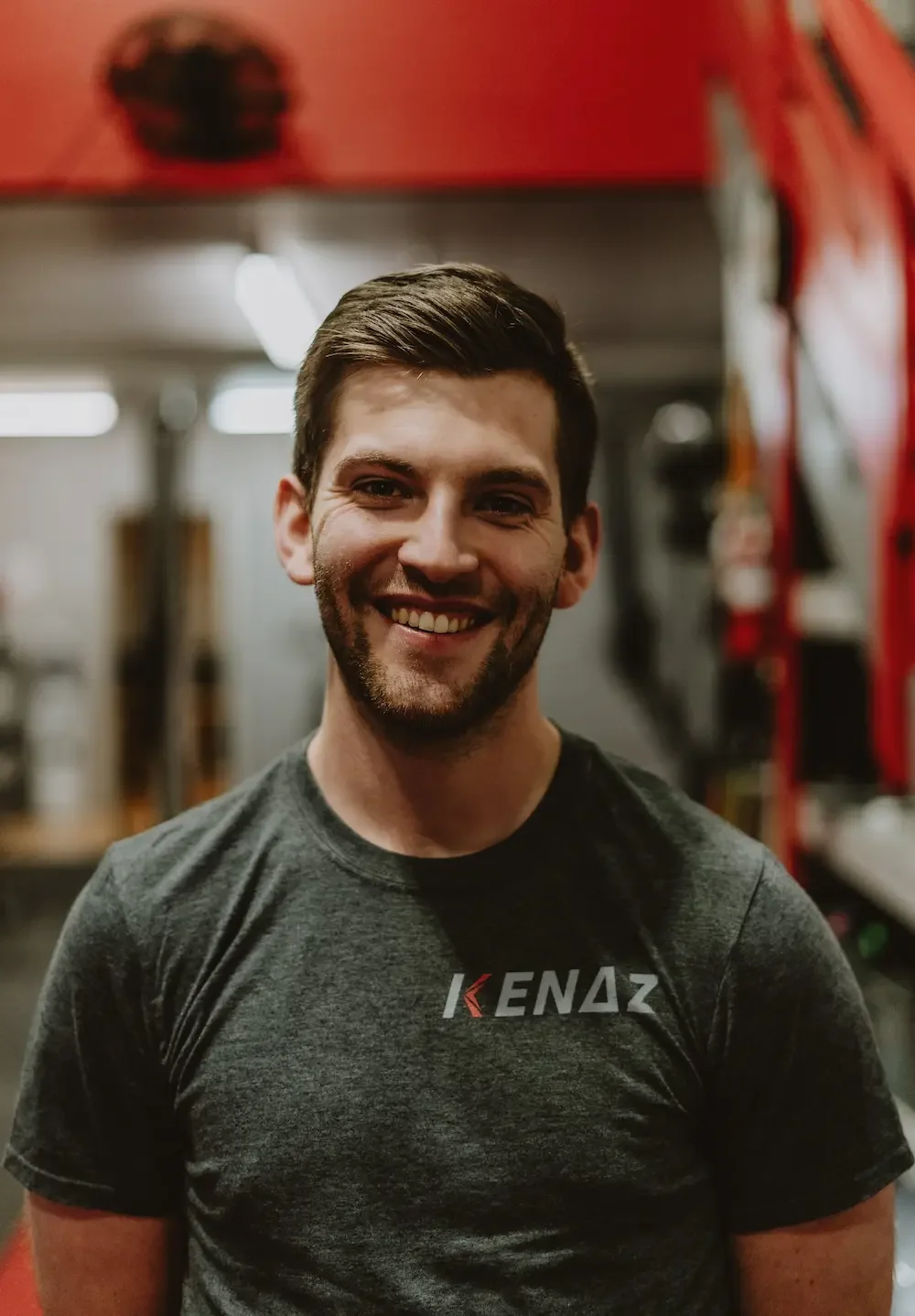 A young man with dark hair and a beard smiling in a gym or fitness center, wearing a dark gray t-shirt with 'KEDAZ' logo.